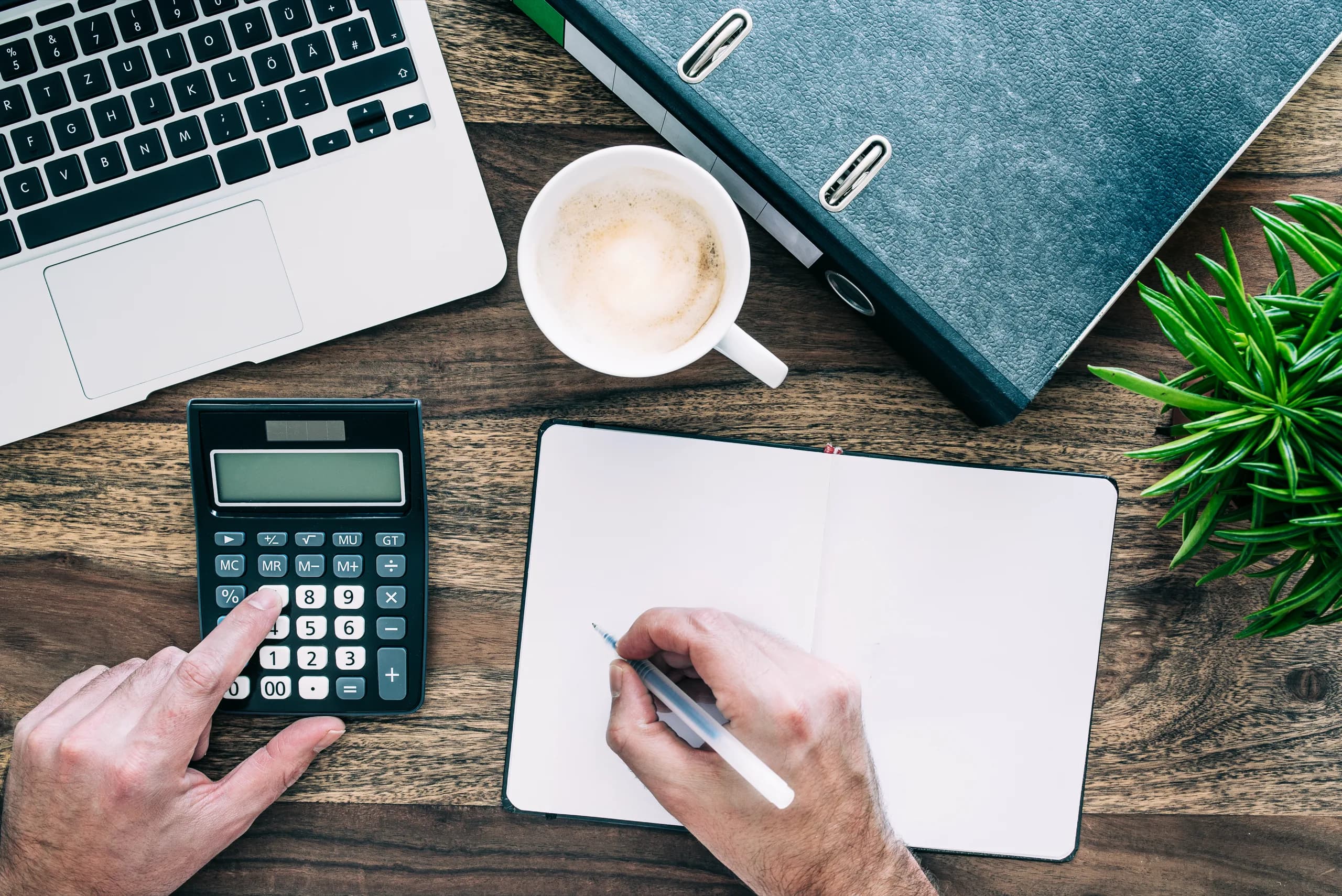 Overhead view of hands with a calculator, notebook, laptop and coffee